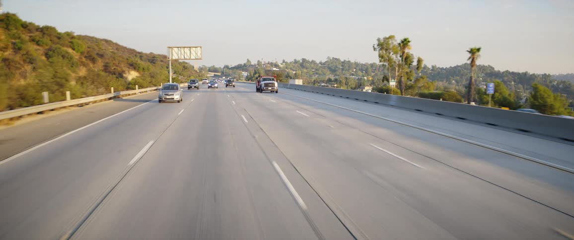 Vehicles traveling on a highway in a hilly region with lots of trees and buidlings