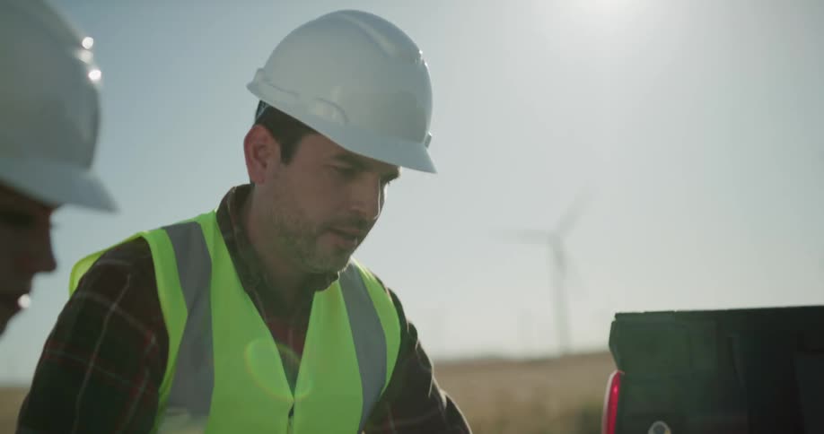 A man and woman in safety vests and hardhats look at a blueprint spread in the bed of a pickup truck parked on a wind farm during the day