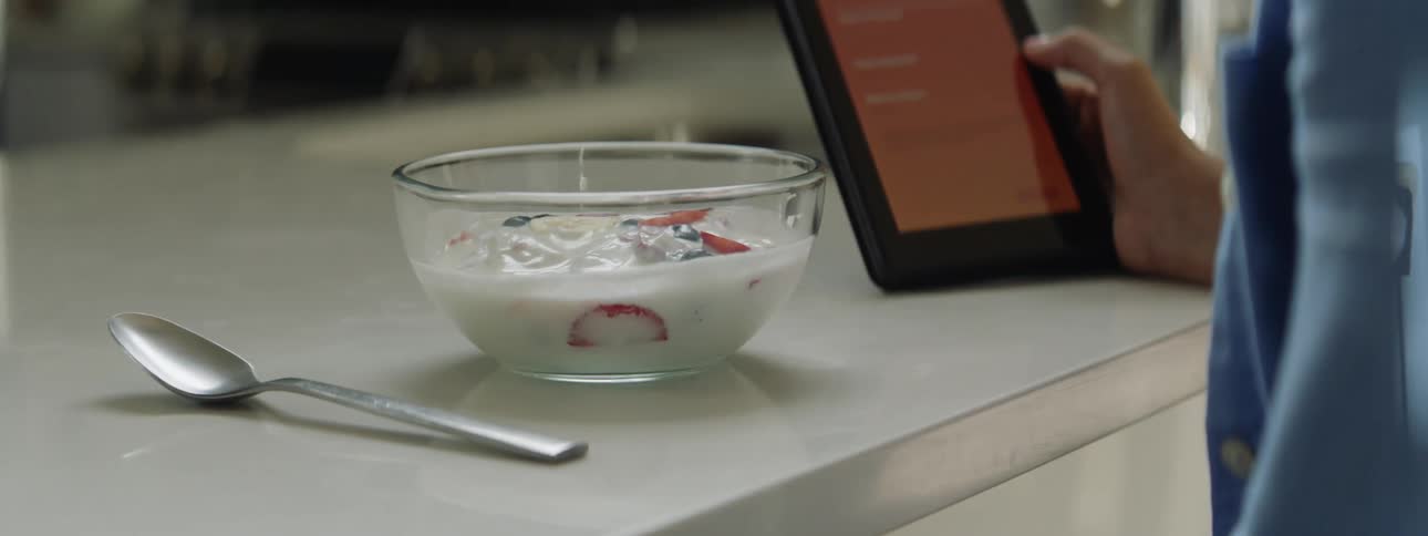 A person sits at a home with a bowl of food in front of them and looks at their tablet