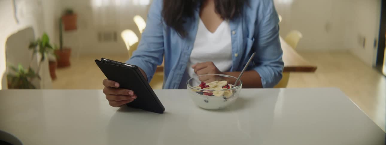 A woman sits at a counter with a bowl of cereal and tablet