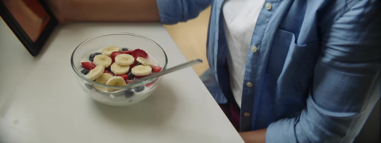 A woman sits at her table and looks at her tablet while eating a bowl of yogurt with fruit