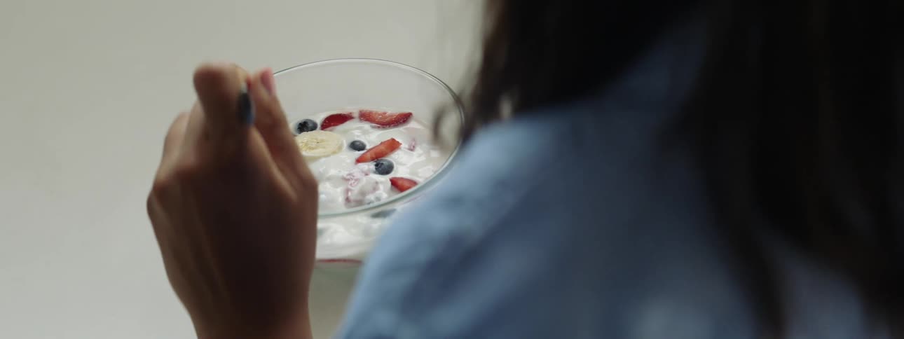 A woman dips a spoon into a glass bowl of yogurt and sliced fruit before lifting it towards her mouth
