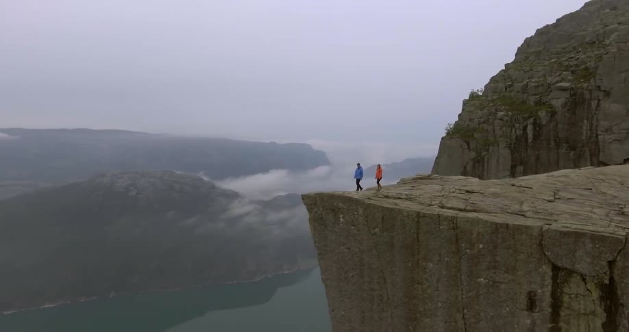 Two people walk to the cliffs edge overlooking a mountain lake