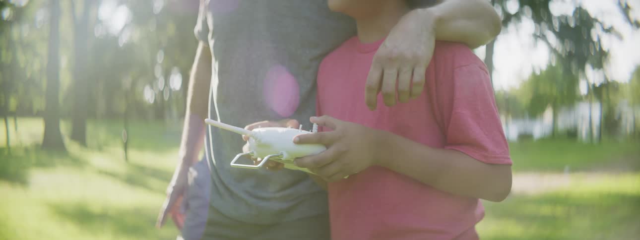 A man drapes his arm around his sons shoulder as they play with a remote control toy outdoors