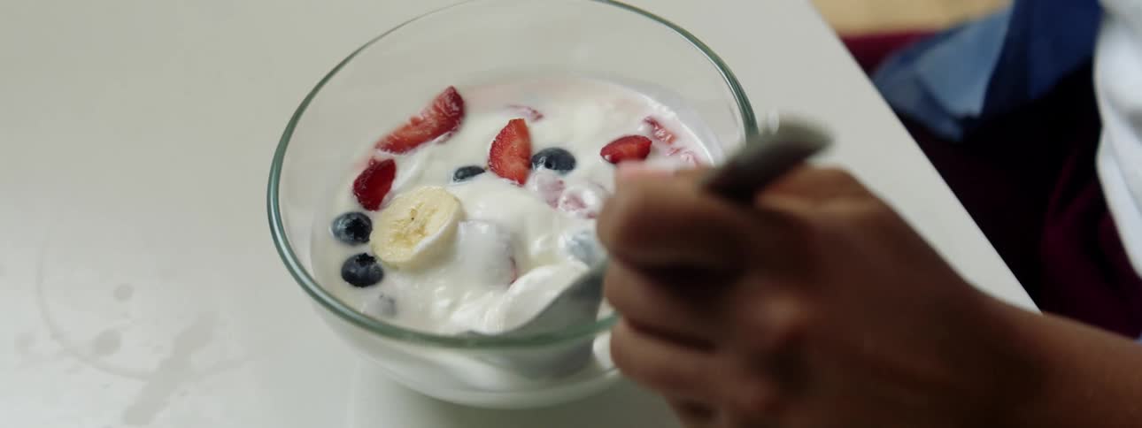 A young woman picks up a spoon from the table and starts eating a bowl of yogurt and fruit