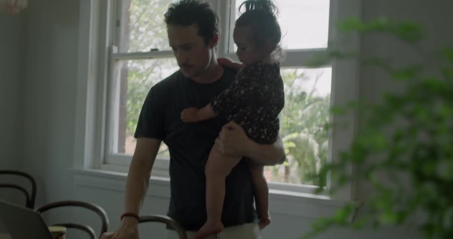 A man carrying a little girl with one arm while standing in a house takes a sip from a tea cup with his free hand before typing on a laptop on the table before him