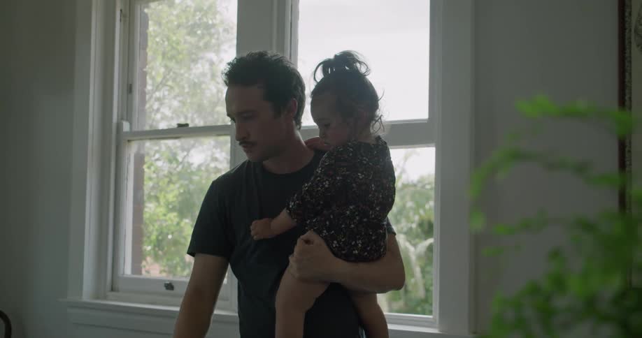 A man holds his toddler daughter while trying to work on his laptop
