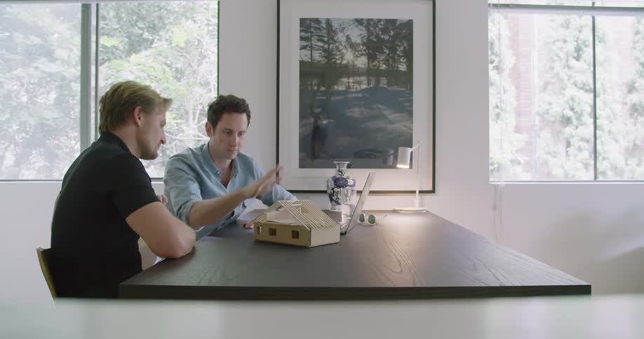Two men in an office look at a model of a building and a laptop as they work together