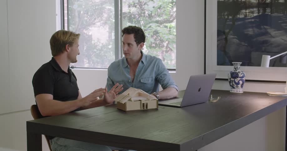 A pair of men sitting together at a table speak to one another while looking at a model house made of cardboard