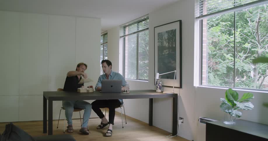 Two men sit at a desk and look at a model of a building as they work