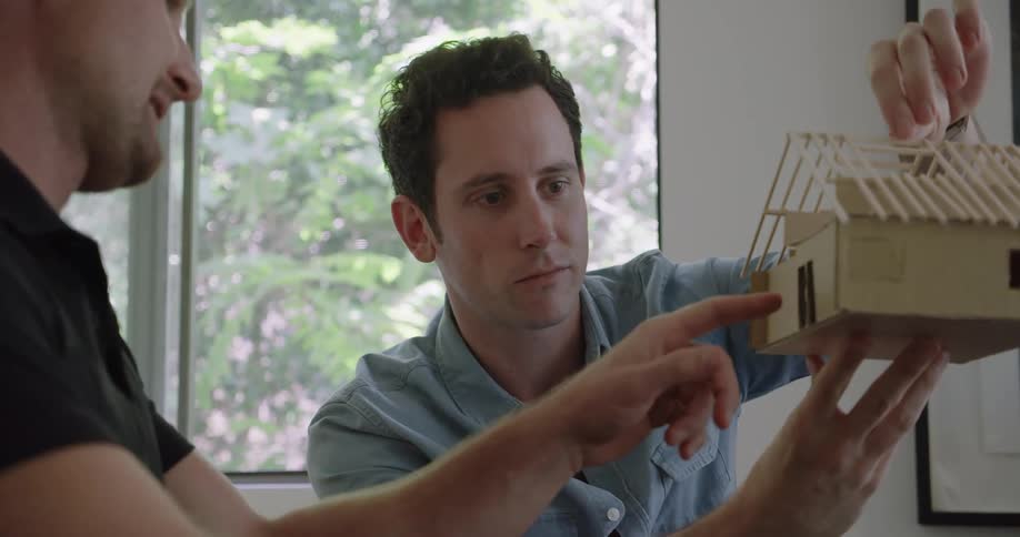 A boss and his employee discuss a model of a house while sitting indoors