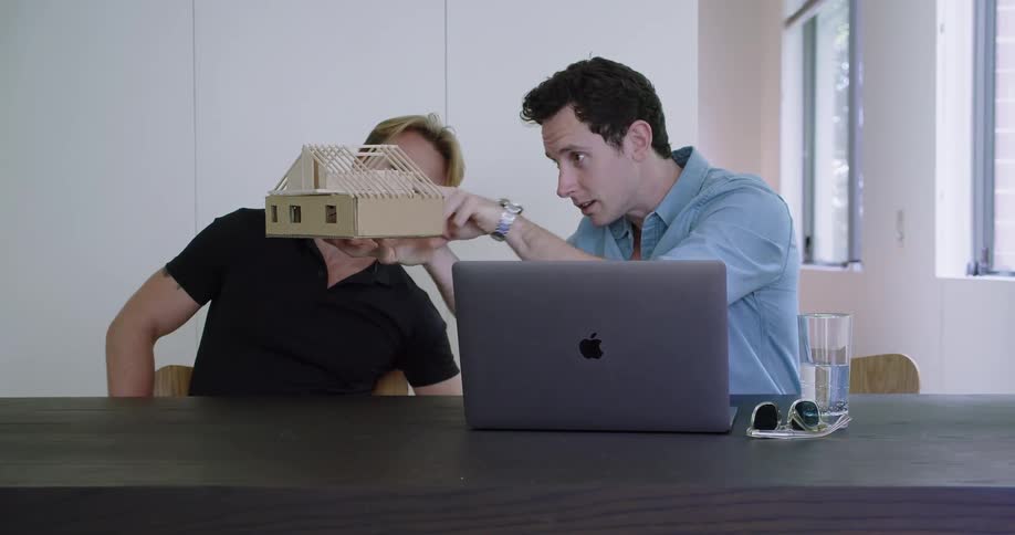 Two men sitting at a table looking at a miniature house and a laptop