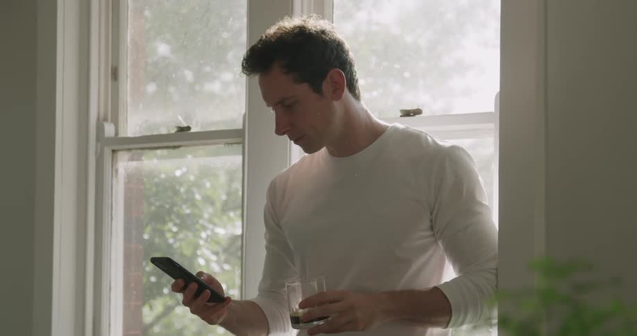 A man stands by a window scrolling through a cell phone and taking a drink from a glass