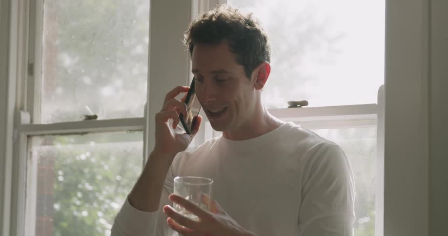 A man holds a glass in one hand and speaks into a phone as he stands indoors in front of a window