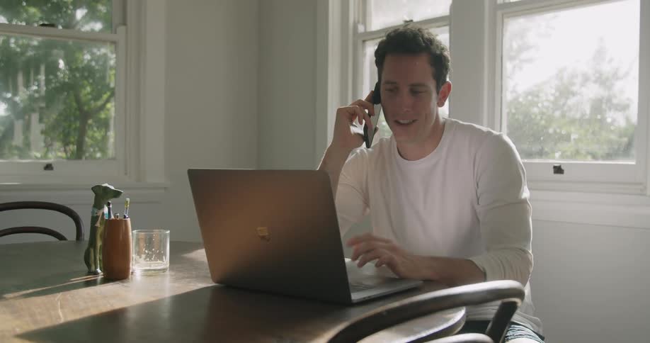 Man sitting talking on cell phone while scrolling on laptop indoors