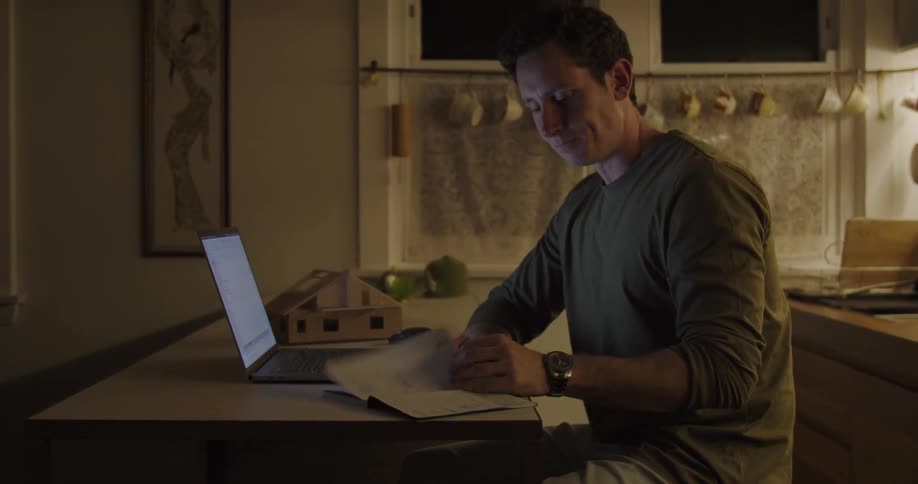 A man in a room sitting at a desk looking at a book and typing on a laptop