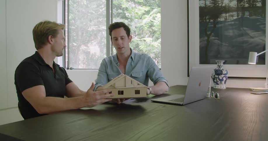 Two men sit at a large office table holding a model building structure and talking about it together