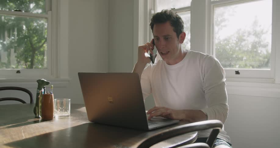 Man talking on a smartphone while working on a laptop at home in the morning