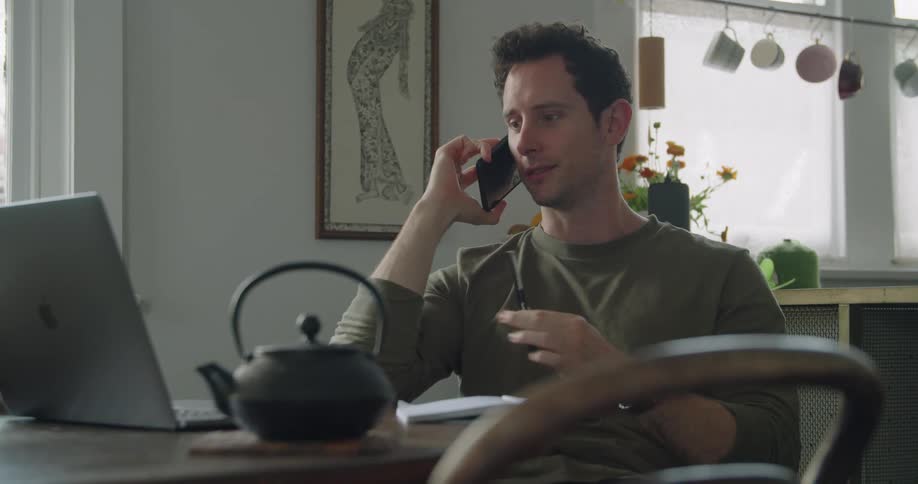 A man smiles as he talks on his smartphone while sitting at his kitchen table in front of his laptop