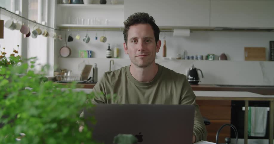 A man is sitting in a kitchen at a table with a laptop in front of him and smiling at the camera