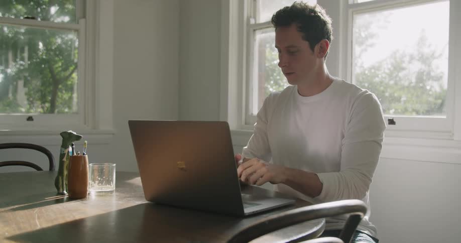 A man sits at a table at home while talking on a phone and working on a laptop during the day