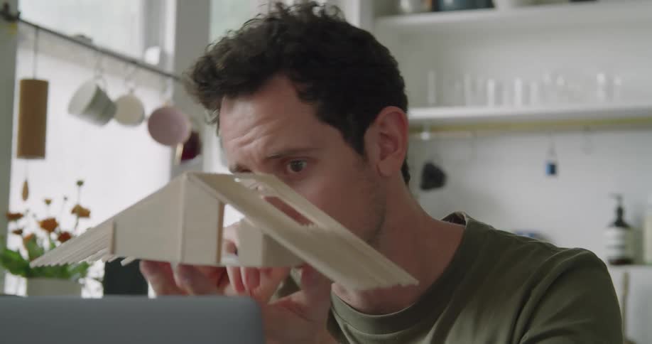 A man holds a small model building structure and appears to be cutting or marking it
