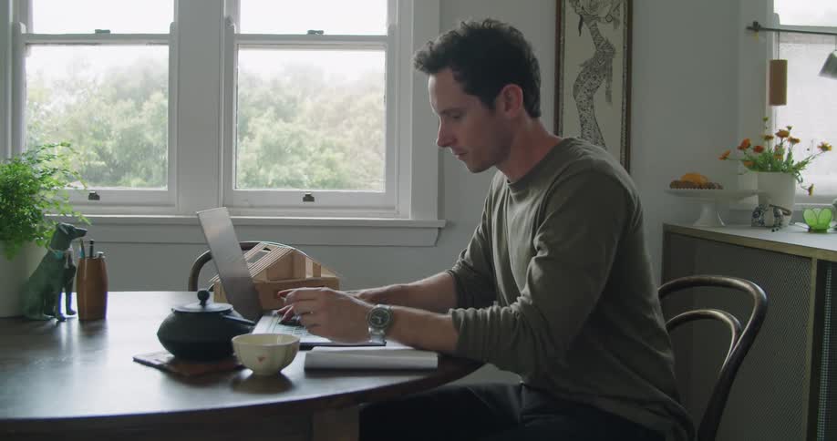 A man sits at a table at home typing on a laptop during the day