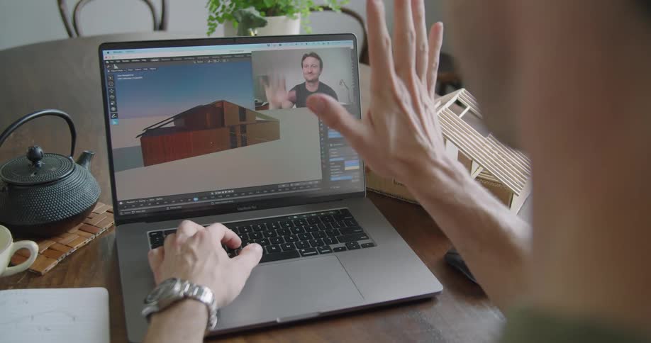 A man uses his laptop to wave at another man through a video conference who returns the greeting
