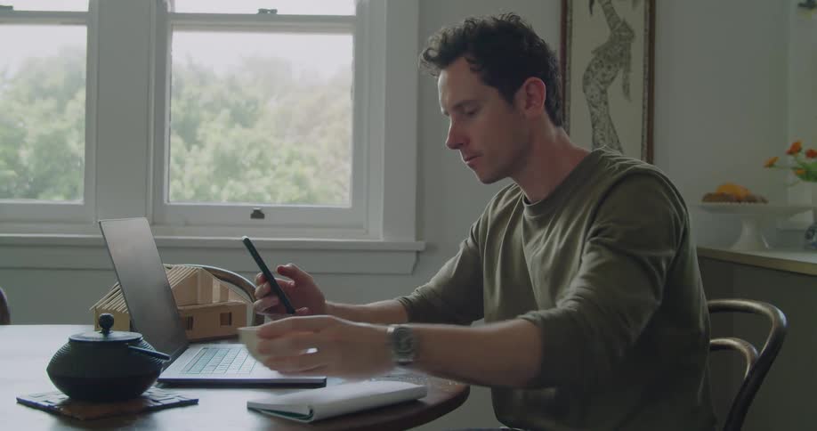 A man with a smart phone sitting on a chair by a table next to a laptop