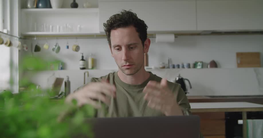 A man is sitting in his kitchen by a laptop