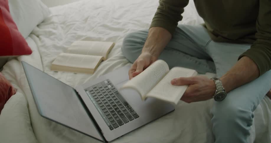 A man looking at a book by a laptop