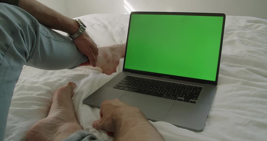 A man sits on a bed looking at a computer with a green screen
