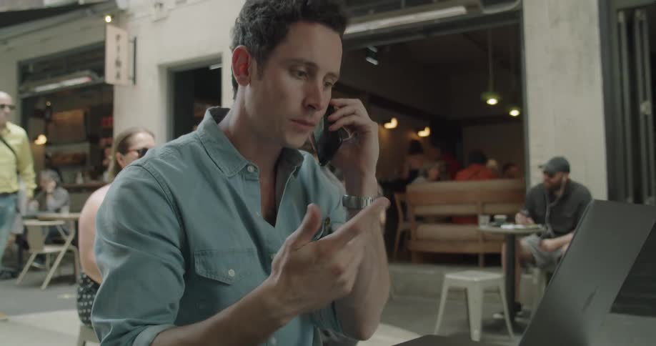 A man sits at a cafe outdoors talking on the phone and typing on his laptop