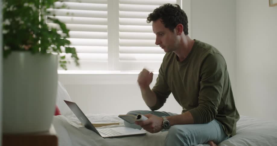 A man in a room sitting on a bed looking at a book and using a laptop