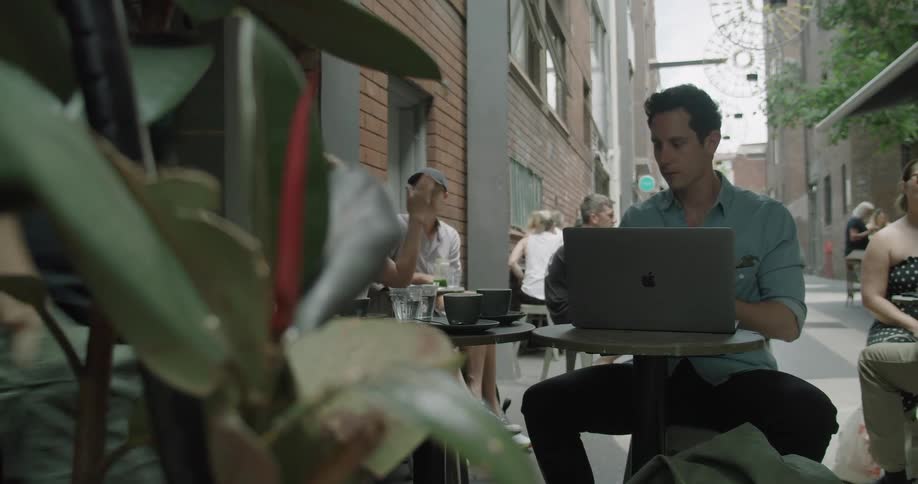 A man drinks from a mug while using a laptop at an outdoor table at a cafe during the day