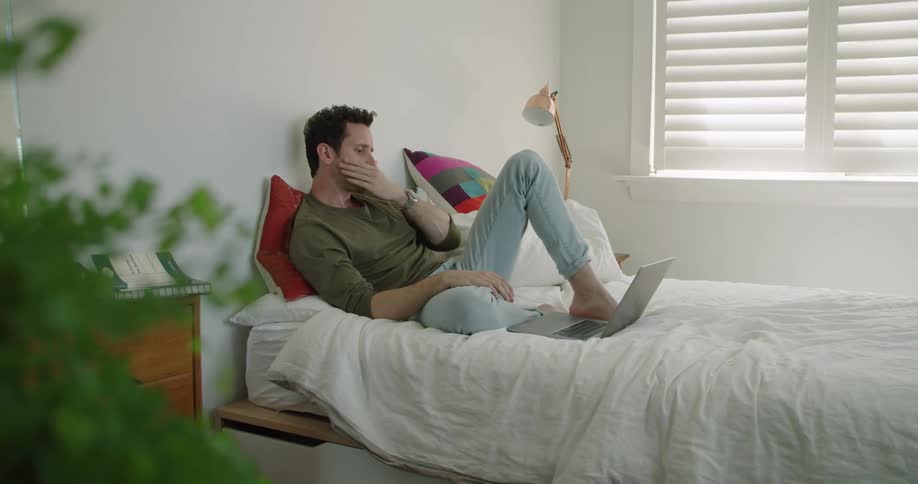A man reclines on his bed while looking at his laptop screen in front of him