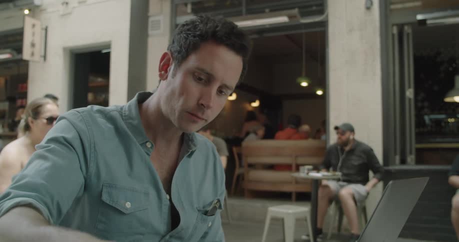 A man sips on a small mug in a cafe and then types on the laptop in front of him