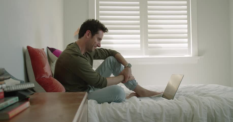 A man sits on his bed at home and video chats with someone on his laptop