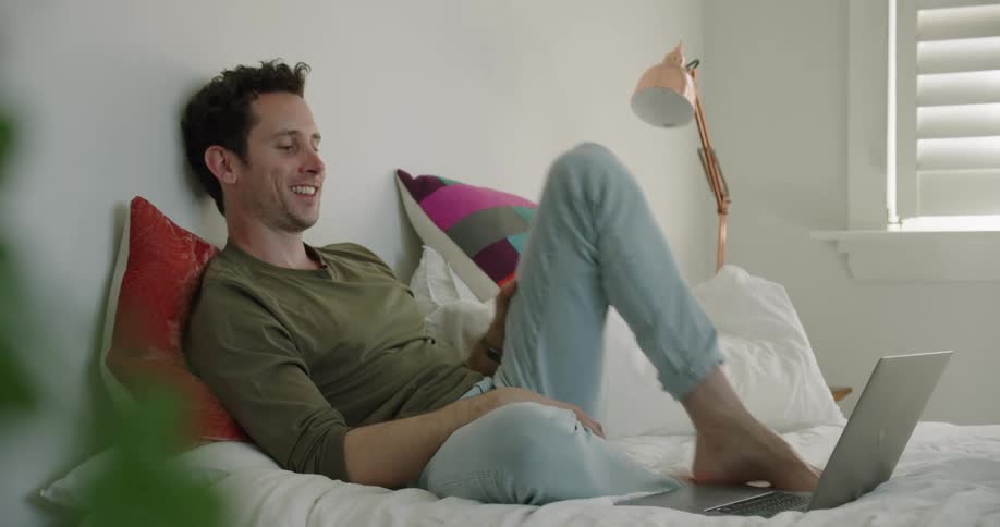 A man resting on a bed laughs while watching the screen of a laptop during the day