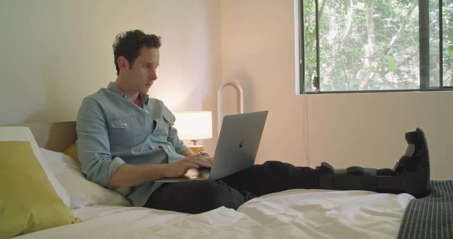 A man with a broken leg adjusts himself as he lies in bed and types on his laptop