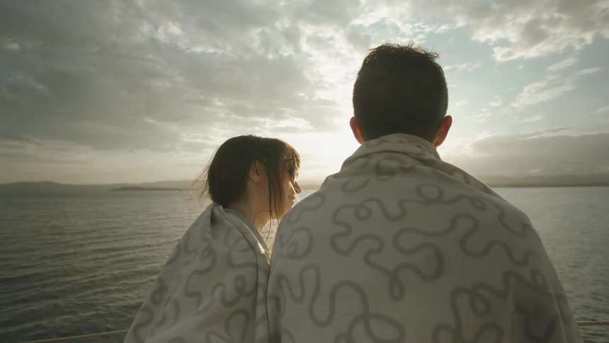 Man and woman in blankets talking next to a lake while the sun sets