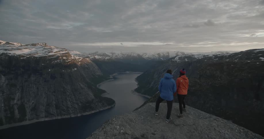 A man and woman stand on a cliff's edge overlooking a vast mountain valley on a cloudy day