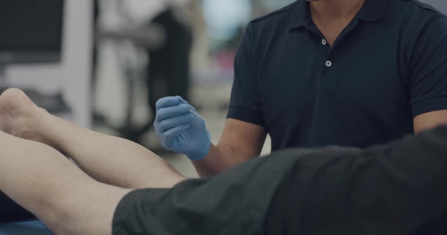 A man sits next to a person laying on stomach and holds a needle in his gloved hand