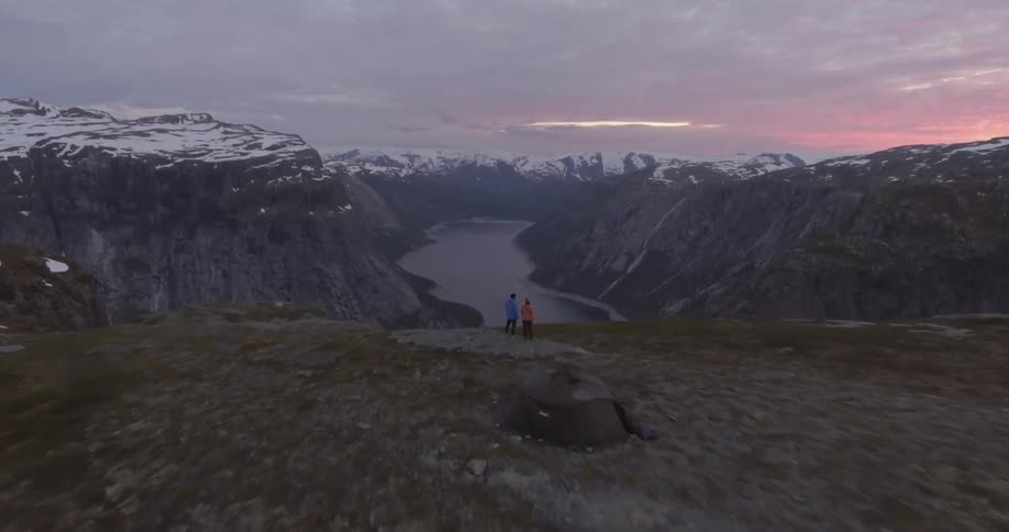 Two people standing on a hill overlooking a river in between snow capped mountains