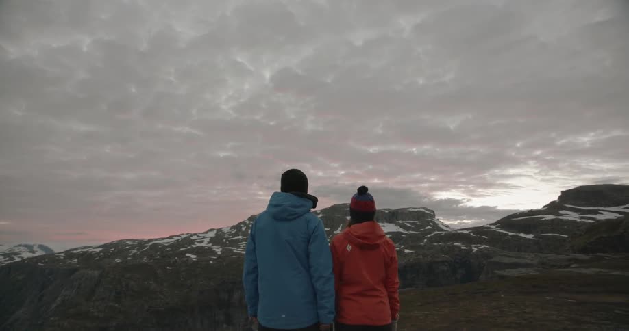 Two people are standing on a cloudy day looking at mountain tops