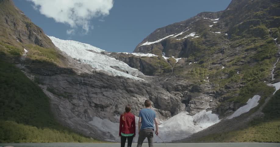 A man and woman stand on the edge of a lake looking up at a mountain with snow on it