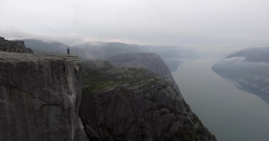 A man and woman stand on a cliff's edge overlooking a vast river valley on a cloudy day