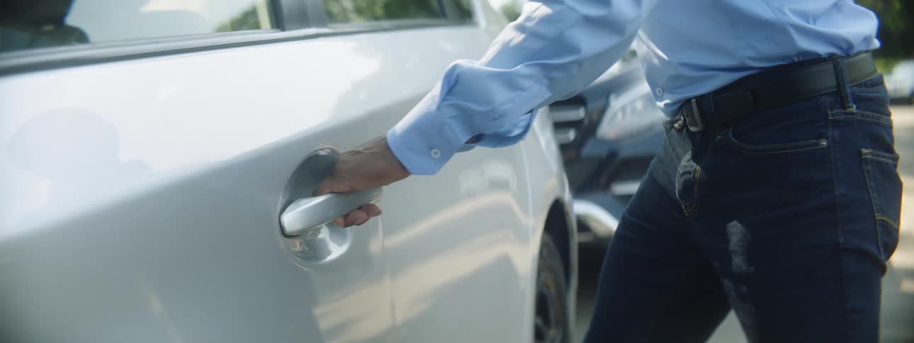 A man wearing a blue dress shirt and jeans opens the car door with his hand outside on a street
