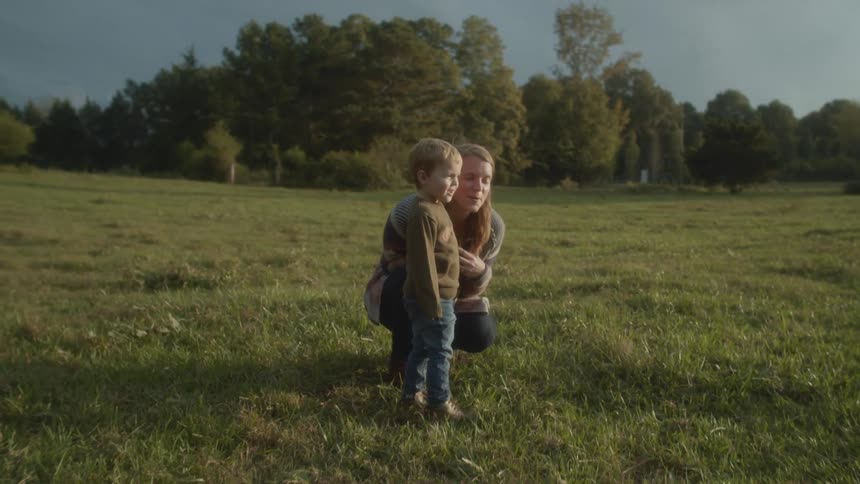 Mother kneels in grass talking to young son who is standing with trees in the back ground