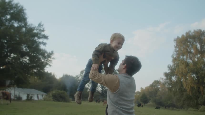 A man holds his toddler son above his head and spins around in circles in a rural yard